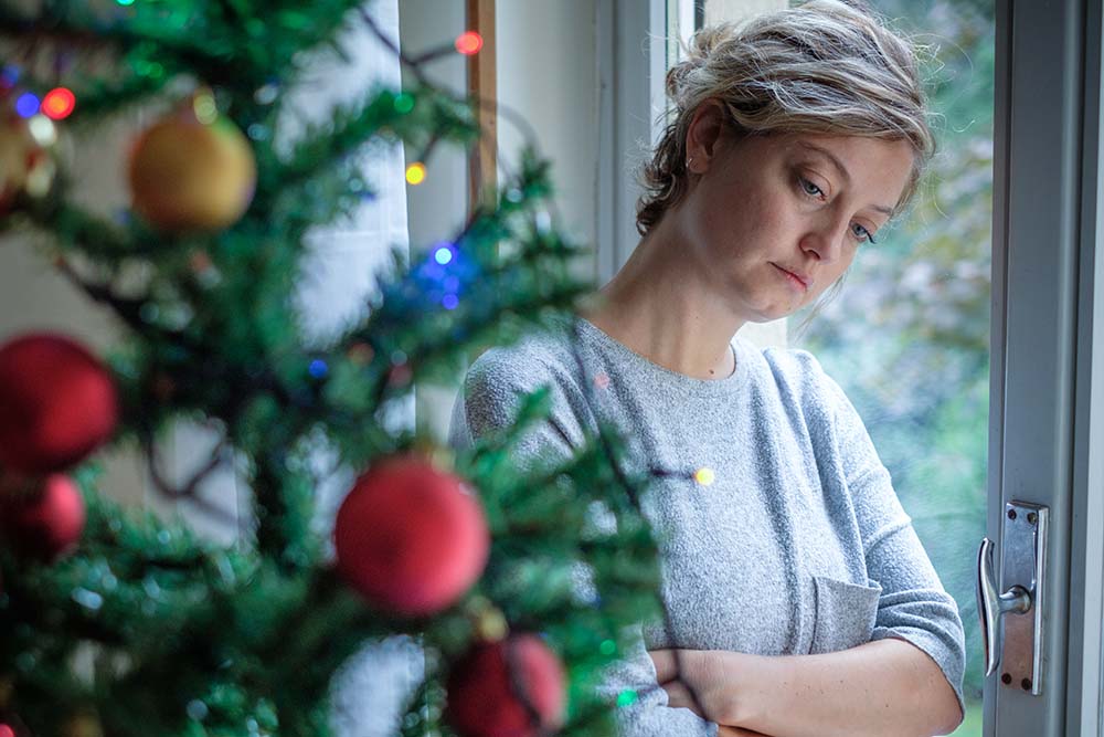 Woman with short hair crosses her arms across her chest and leans her head against a window. She is standing near a Christmas tree.