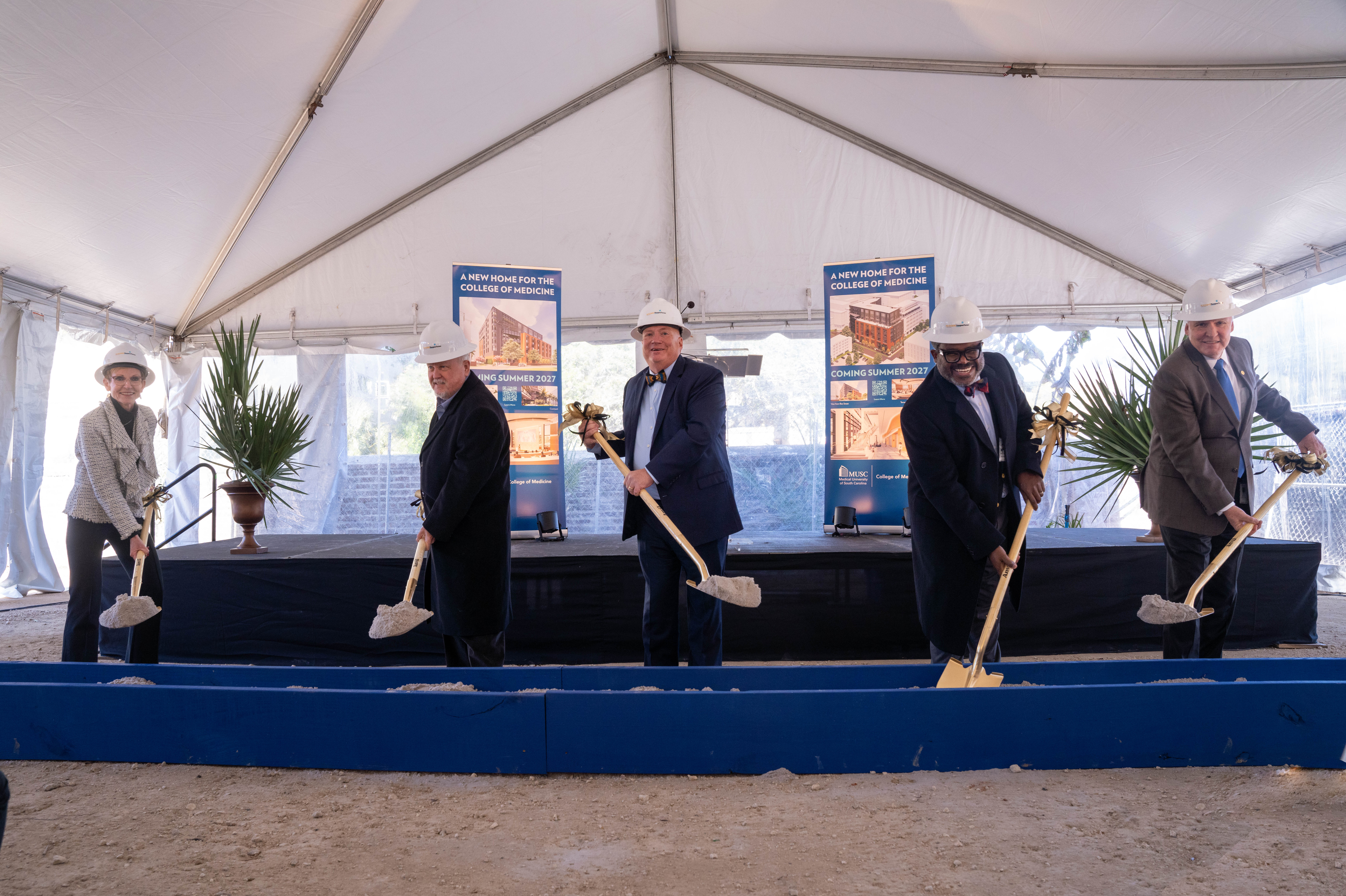 Five people wearing hard hats and holding shovels lift dirt from a blue trough. They are in a white tent.