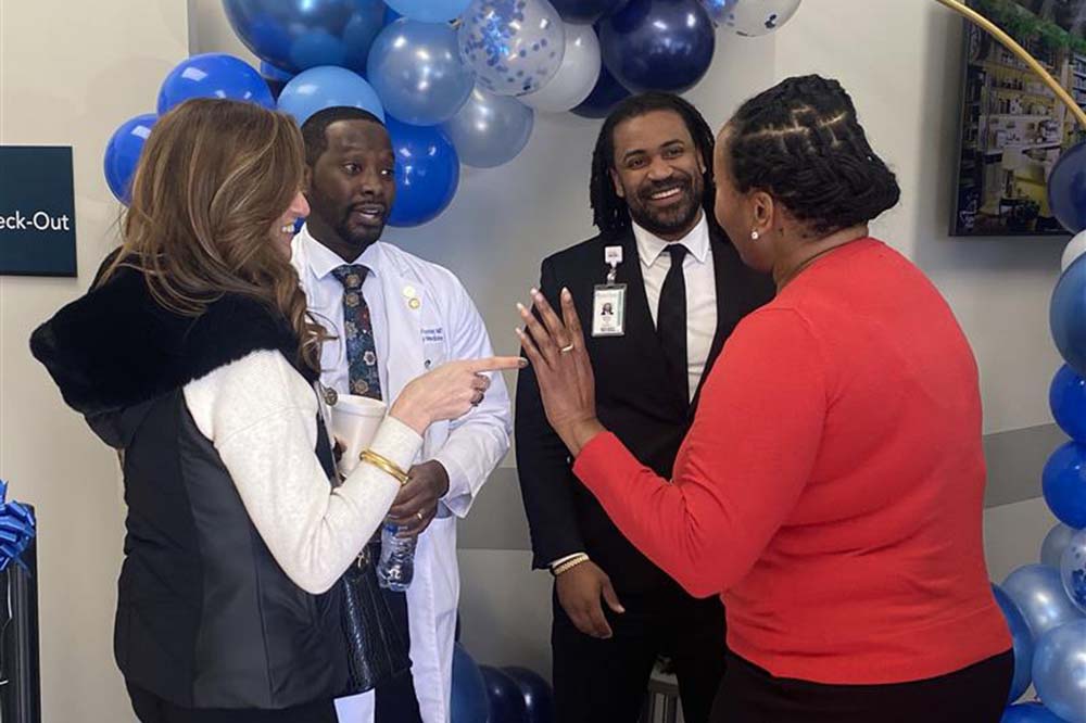 Four people smile and talk in front of blue helium balloons.