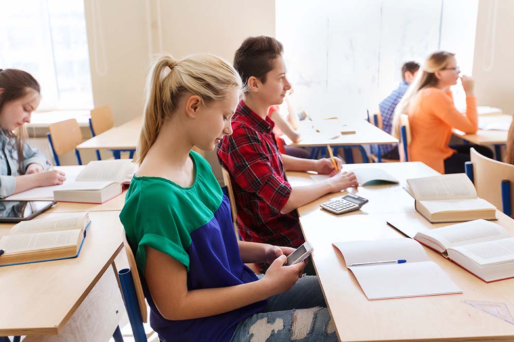 Girl with blond ponytail wearing a green and blue top looks at a cellphone under her desk in a classroom.