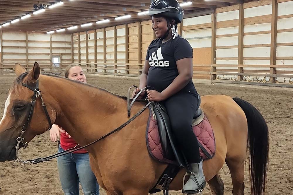 A very happy looking girl wearing a black t-shirt and pants and a black helmet sits on a brown colored horse.
