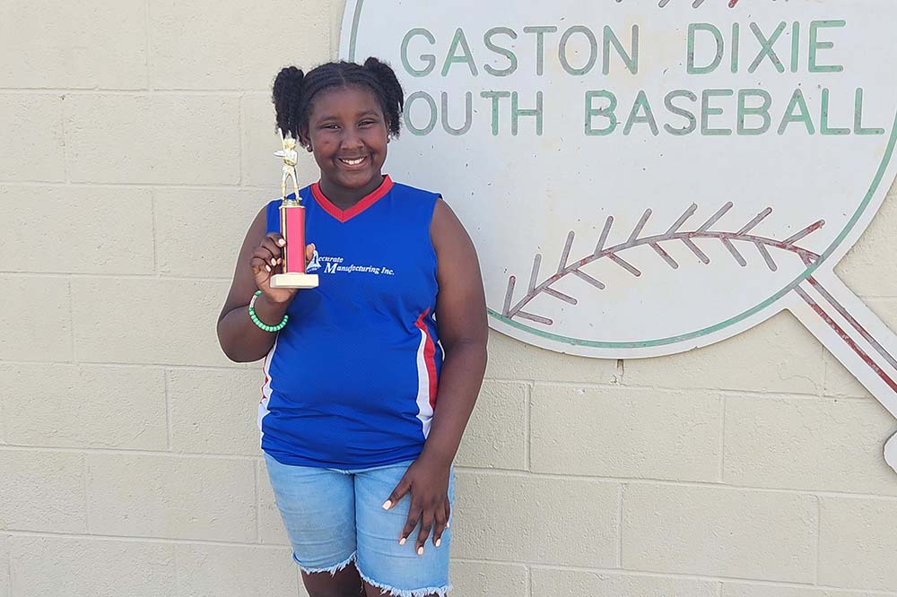 A smiling girl wearing a blue top and shorts holds trophy in front of a sign that says Gaston Dixie Youth Baseball.
