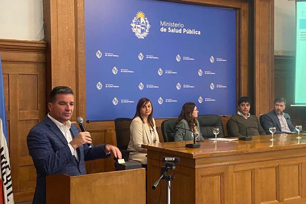 A man in a suit coat and white shirt holds a microphone while speaking behind a podium. Four people are seated at a table to his left. A screen behind them says Minister de Salud Public.