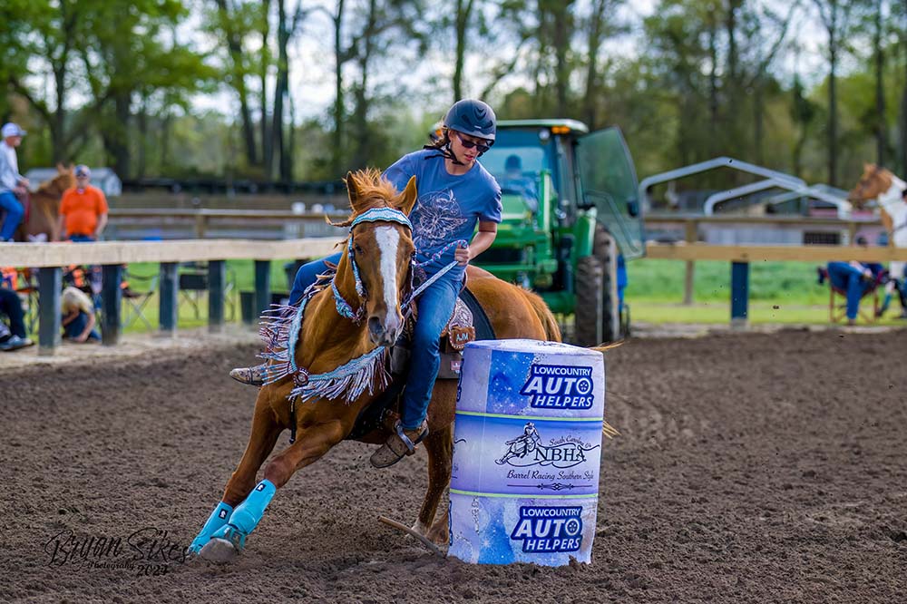 Girl wearing a riding helmet turns a brown horse around a blue barrel.