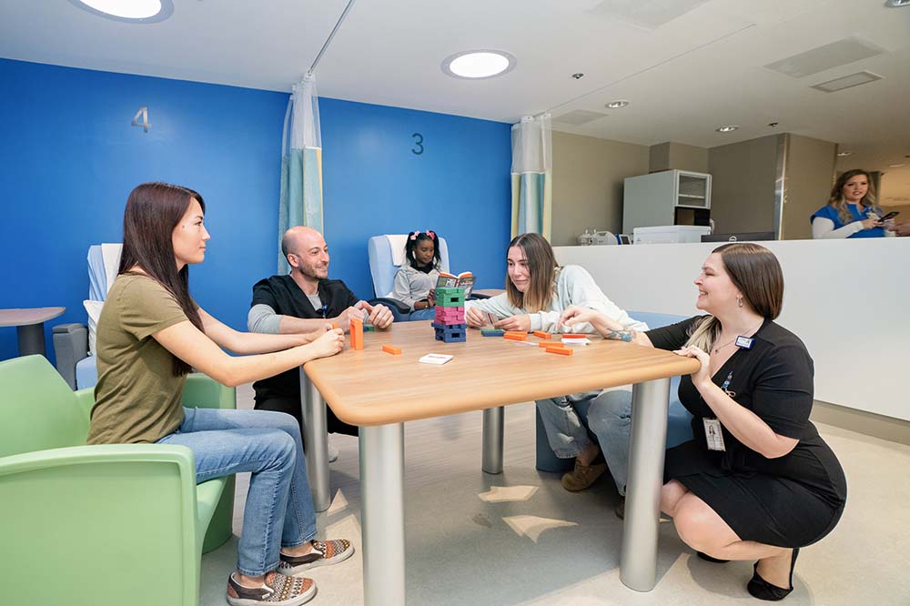 Four people around a small table with blocks in the middle. A young women in a chair behind them is reading.