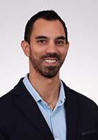 Headshot of man with dark hair and a beard. He is wearing a navy jacket and lighter blue collared shirt.