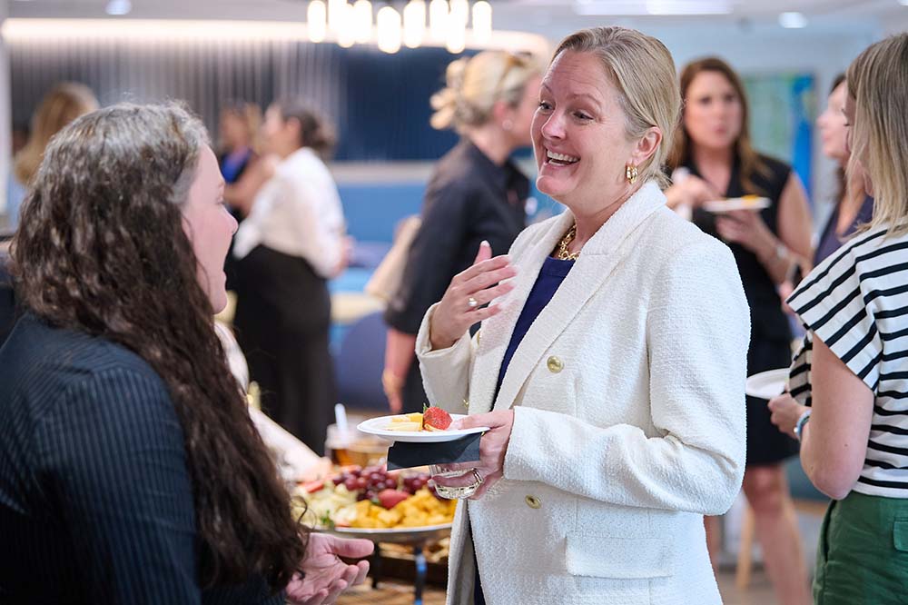 A smiling woman wearing a white coat talks to another woman while holding a plate at what looks like a reception.
