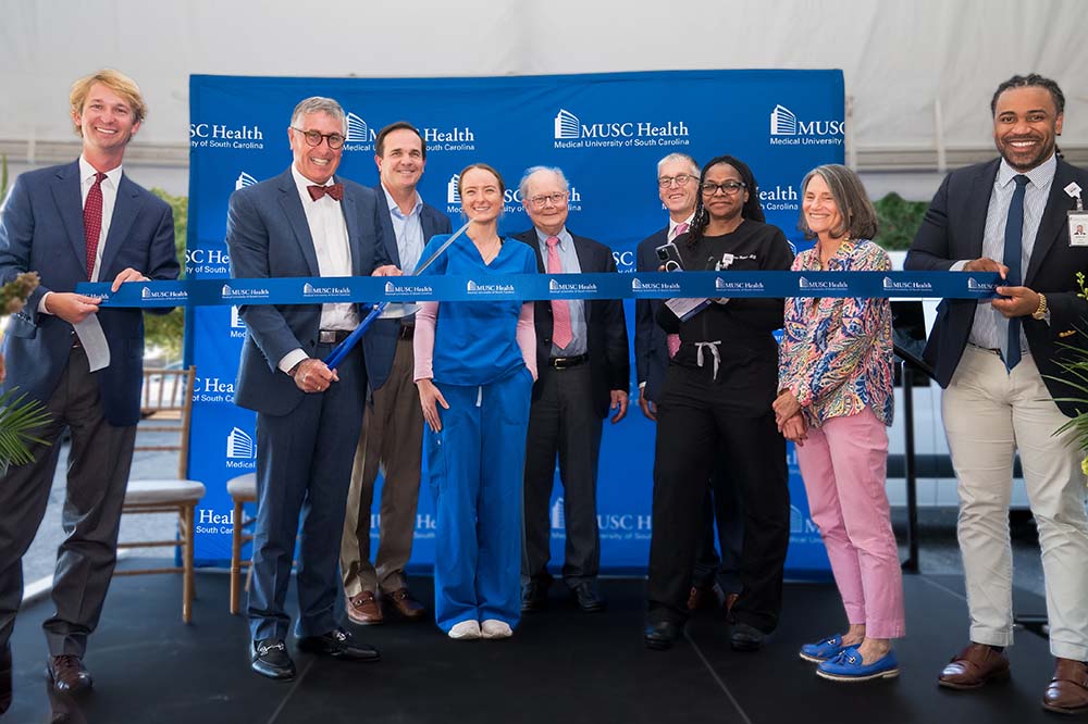 A group of men and woman in front of a blue backdrop that says MUSC Health pose as they prepare to cut a large blue ribbon.
