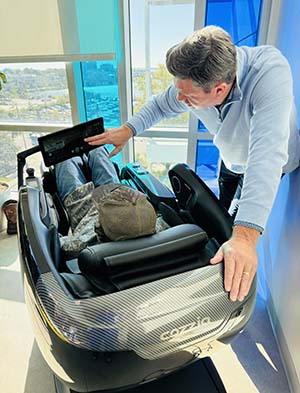 A person is seated in an elaborate looking chair with a small screen. A man stands beside it, pointing to the screen.